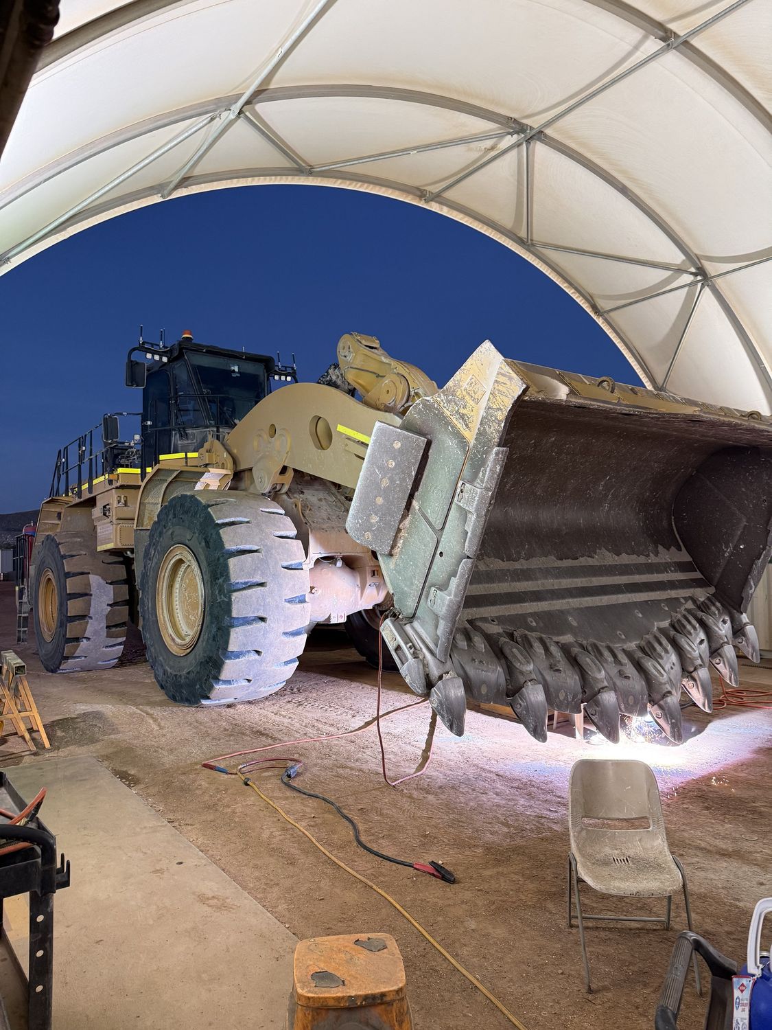 Boilermaker welding the cutting edge of a raised wheel loader bucket at night on a mine site